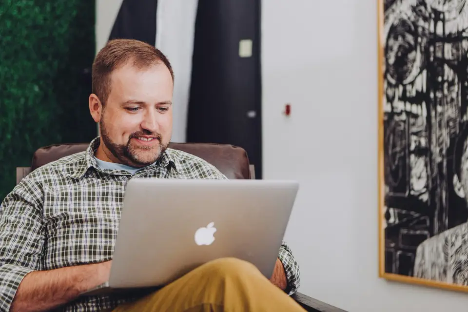 A business owner in a checked shirt smiling as he works on a laptop in a relaxed office